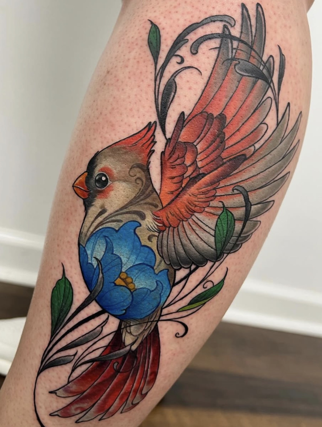Neo-traditional female cardinal with spread red wings, swirling filigree feathers and a blue bloom, on the calf